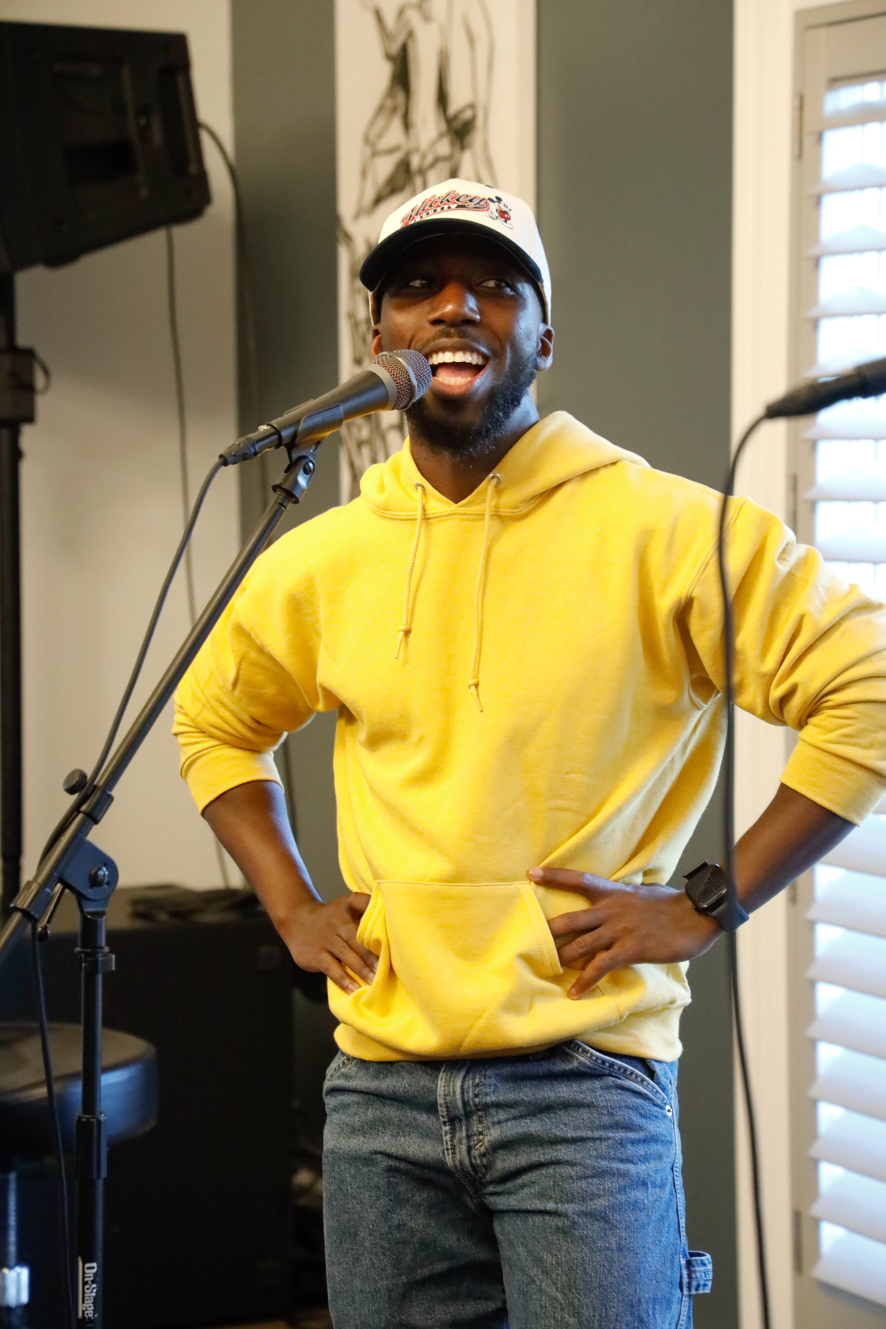 Performer in a yellow hoodie and Mickey cap smiling on stage at the microphone
