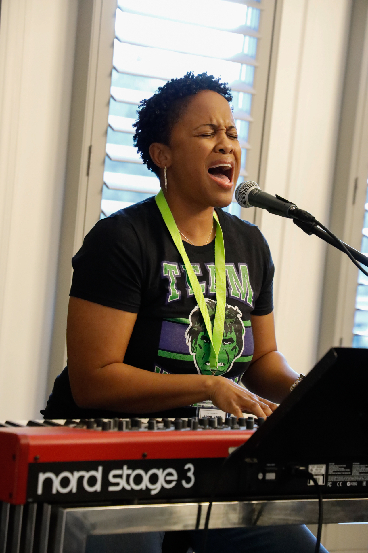 Vocalist belting into a microphone while playing a Nord Stage 3 keyboard