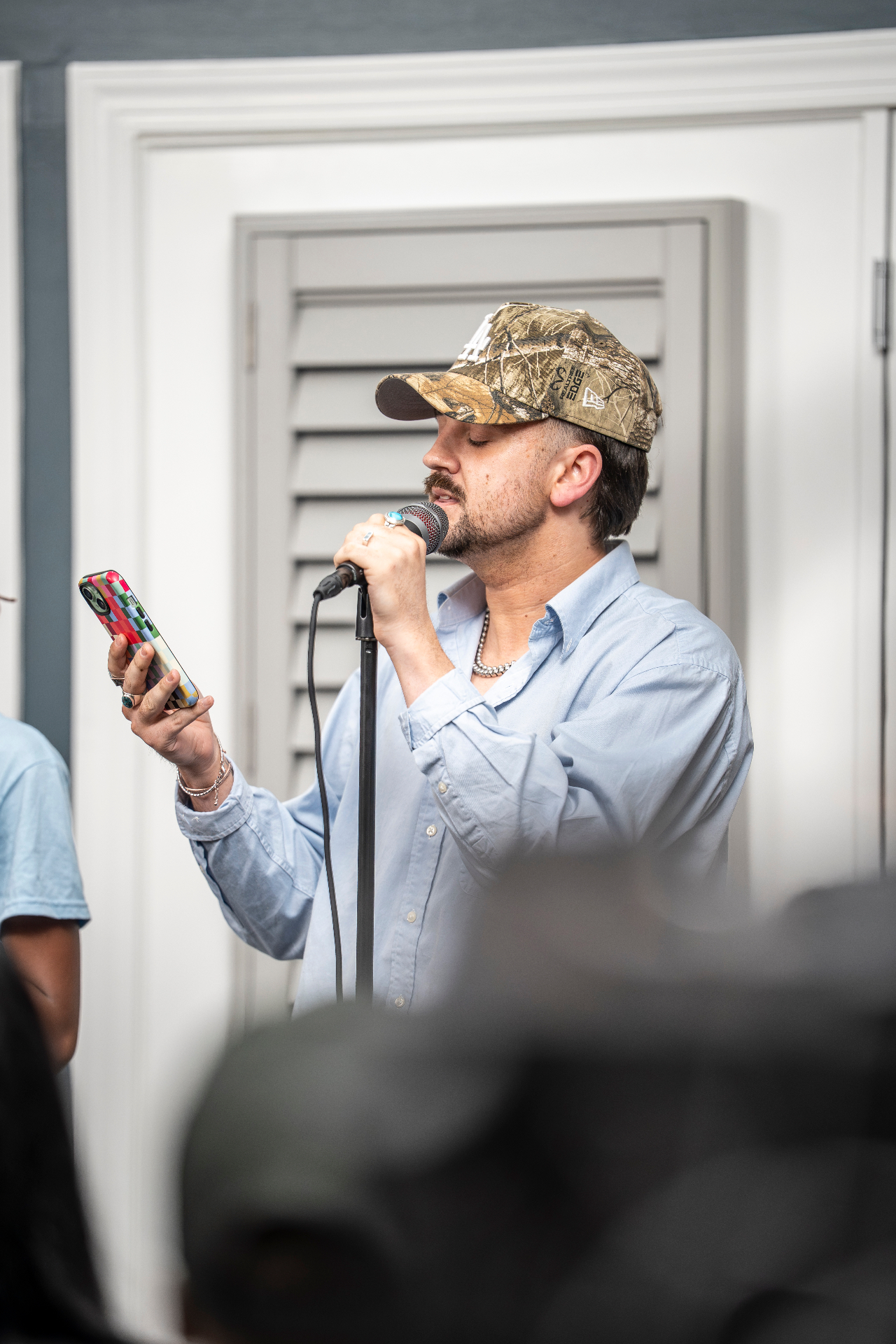 Performer in a camo cap singing into a microphone while reading lyrics from a phone